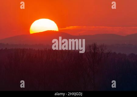 The top of the mountain is hidden in the twilight. Behind the mountain you can see. partly behind it, a red shield of the setting sun is hidden. Stock Photo