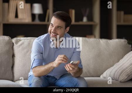 Smiling young male sitting on sofa holding phone looking aside Stock Photo