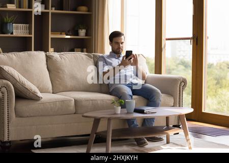 Millennial man wearing wireless headphones relaxing on sofa using phone Stock Photo