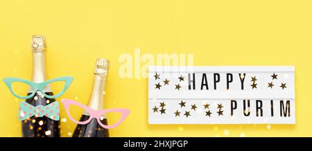 Purim background. Happy Purim written in lightbox, party props, masks, and champagne on a bright yellow background. Top view, copy space. Banner Stock Photo