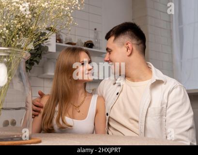 Young happy couple smiling and looking tenderly at eyes. Beautiful woman and handsome man sitting at table in white modern kitchen in Scandi style. Gentle relations and love concept. High quality photo Stock Photo