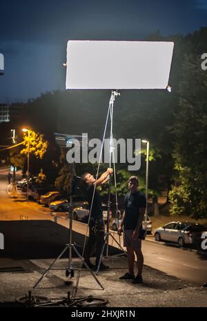 Behind the scene. Filmmaking lighting technician electric engineer adjusting and setup lights for movie film scene outdoor by night. Light Department. Stock Photo