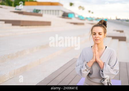 Medium shot of relaxed Caucasian young woman practicing yoga performing namaste pose with closed eyes outside in city park. Stock Photo