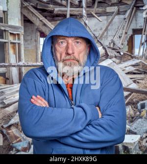portrait of sad and gloomy senior man in a hoodie against rubbish of a ruined house Stock Photo