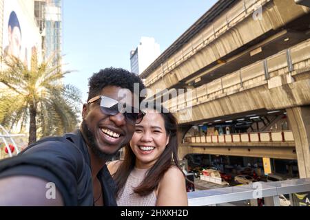 Two multethic smiling friends embracing while taking a selfie outdoors Stock Photo