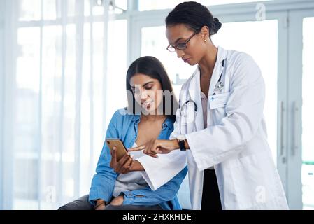 Ill send you some helpful health tips to you. Shot of a young doctor using a smartphone during a consultation with her patient. Stock Photo