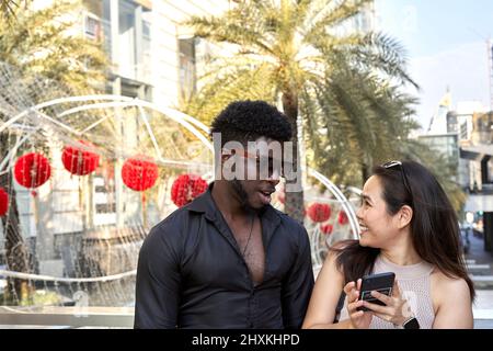 Multiethnic friends chatting while using the mobile outside a mall in summer Stock Photo