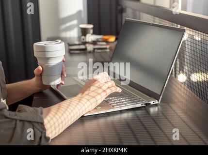 Woman working at laptop computer in coffee shop by window with day light and eco coffee cup. Stock Photo