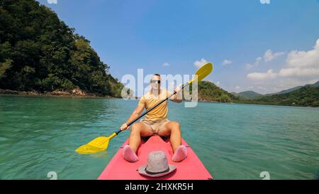 Young man with sunglasses and hat rows pink plastic canoe along sea against green hilly islands with wild jungles. Traveling to tropical countries. Strong guy is sailing on kayak in ocean, front view. Stock Photo
