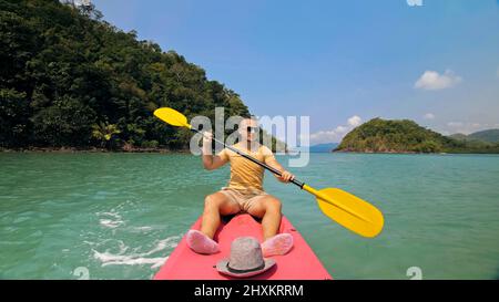 Young man with sunglasses and hat rows pink plastic canoe along sea against green hilly islands with wild jungles. Traveling to tropical countries. Strong guy is sailing on kayak in ocean, front view. Stock Photo