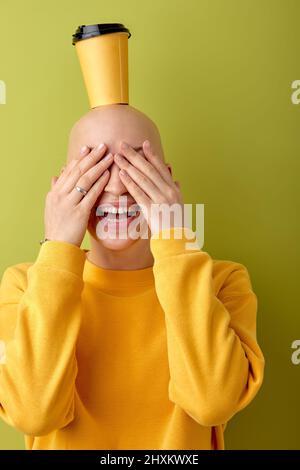 Funny hairless female have fun holding cup of coffee on bald head, enjoy life. Caucasian woman with allopecia disease laughing, dressed in casual yell Stock Photo