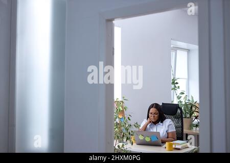 Pleasant black woman sit alone in office behind desk working online on laptop, typing on keyboard. beautiful lady in formal wear concentrated and focu Stock Photo