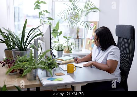 Black Office Female Sitting At Desk Working On Laptop Taking Notes At Work Indoor. Business Schedule Concept. Attractive Lady in White Shirt Concentra Stock Photo