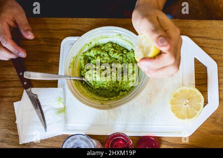 top view of caucasian male hands squeezing a lemon over a bowl preparing guacamole, on a white cutting board on a wooden table in the background Stock Photo