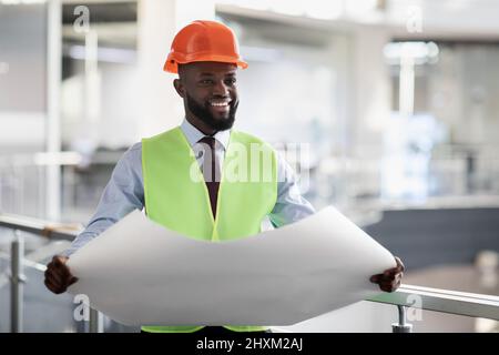 Cheerful black civil engineer in safety workwear holding draft Stock Photo