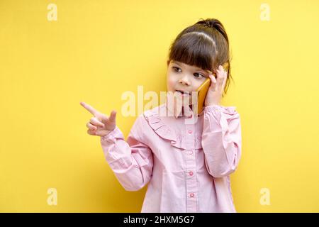 Child girl having serioys talk on phone gesturing, explaining, pointing finger at side. isolated over yellow background Stock Photo