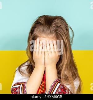 A girl in national Ukrainian clothes, an embroidered shirt, is crying, covering her face with her hands against the background of the Ukrainian flag Stock Photo