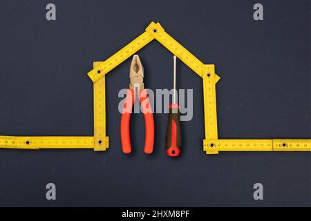 Home repair. Hand tools under house shape yellow wooden measure, black background. Folding ruler and hardware, workshop, construction, renovation conc Stock Photo