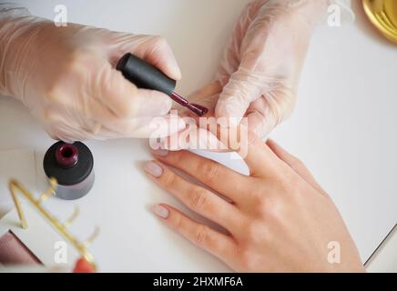 Close up of manicure specialist hands in sterile gloves applying red nail polish on woman fingernail. Professional manicurist doing manicure for female client in beauty salon. Stock Photo