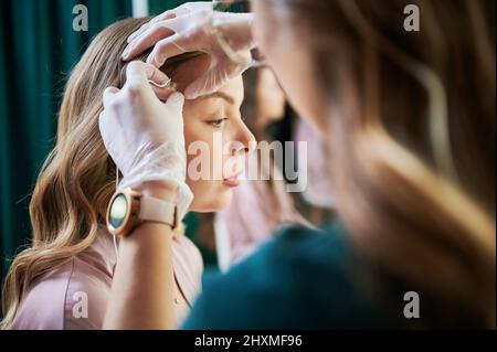 Close up of female stylist in sterile gloves using cosmetic string while correcting woman eyebrows. Young woman having beauty procedure in modern salon. Stock Photo