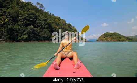 Young man with sunglasses and hat rows pink plastic canoe along sea against green hilly islands with wild jungles. Traveling to tropical countries. Strong guy is sailing on kayak in ocean, front view. Stock Photo