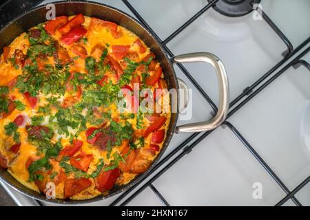 Omelette with vegetables and herbs in black frying pan on white stove. Top view Stock Photo