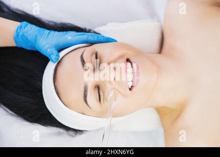 Female cosmetologist doing rejuvenation electric darsonval face procedure in cosmetology clinic to a brunette happy woman. Stock Photo