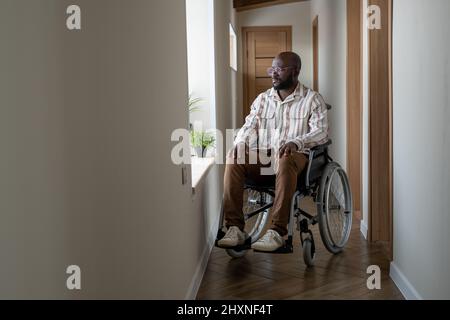 Young African American man with disability looking through window while sitting on wheelchair in corridor of apartment Stock Photo