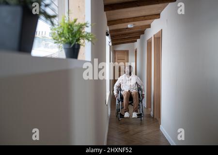 Young man with disability in casualwear moving along corridor of modern apartment while sitting on wheelchair Stock Photo