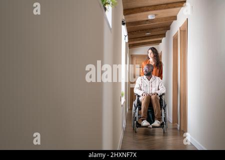 Young black man in casualwear sitting in wheelchair in front of Caucasian female caregiver helping him move in apartment Stock Photo