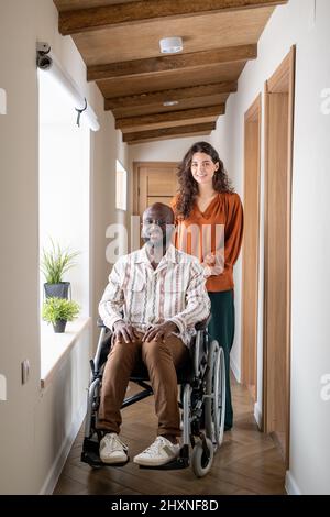Young cheerful man with disability and his caregiver in casualwear looking at camera in corridor of modern apartment Stock Photo