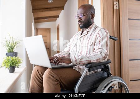 Contemporary young serious black man with laptop on his knees sitting in wheelchair and working in the net at home Stock Photo