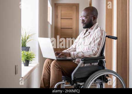 Young serious black man with disability concentrating on network while sitting in wheelchair in corridor of apartment Stock Photo