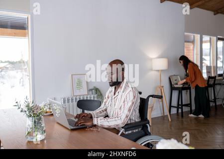 Serious African American man in wheelchair sitting by table in front of laptop and networking against female switching on record player Stock Photo