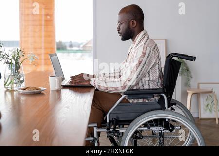 Side view of serious black man working in the net or analyzing online data while sitting in front of laptop by table Stock Photo