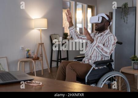 Contemporary software developer in vr headset touching virtual display while sitting in wheelchair and working from home Stock Photo