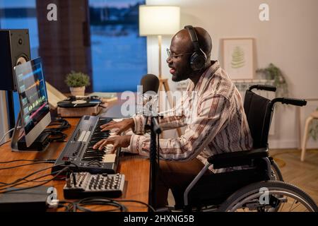Side view of African American musician in wheelchair recording his songs while playing piano keyboard in front of computer by table Stock Photo