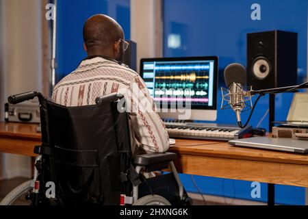 Rear view of man with disability using sound recording program while sitting by table in front of computer monitor in studio Stock Photo