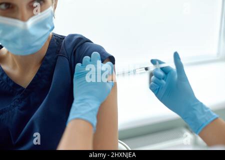 Woman in medical face mask getting injection at hospital. Doctor or nurse preparing syringe to give shot to female patient Stock Photo
