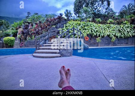 Senior female's legs with red nails in front of swimming pool with tropical plants Stock Photo
