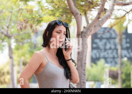 Adult woman with a serious expression talking with her phone outside with a defocused background of trees Stock Photo