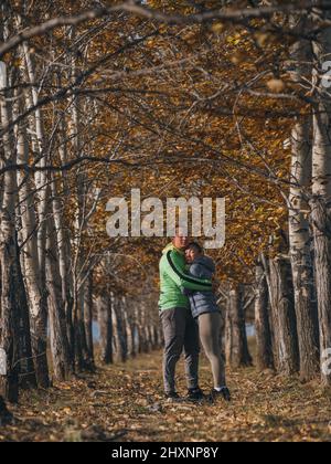 Man and woman in yellow green gray sportswear in the autumn. Lovely couple of travelers hug and kiss in arch of dry trees. Two travelers are walking in the fall. Stock Photo