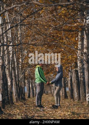 Man and woman in yellow green gray sportswear in the autumn. Lovely couple of travelers hug and kiss in arch of dry trees. Two travelers are walking in the fall. Stock Photo