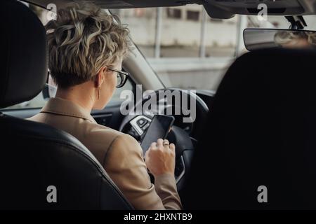Business Woman Using a Smartphone While Driving Car, View from Back Seat Stock Photo