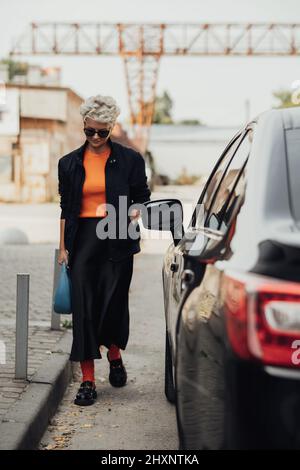 Stylish Young Woman in Jacket and Sunglasses Moving Near the Black Car Stock Photo