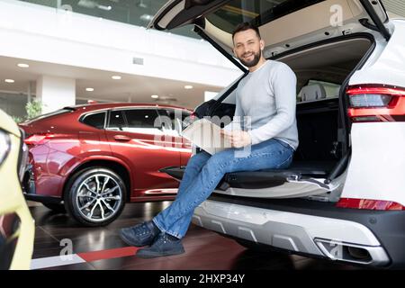a buyer studying a booklet in a showroom of a car dealership sitting in the trunk of a crossover looking at the camera with a smile Stock Photo