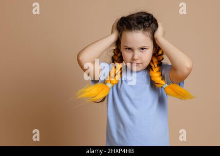 Offended young Caucasian girl touching head with hands looking sullenly at camera having kanekalon braids of yellow color in blue t-shirt on beige Stock Photo