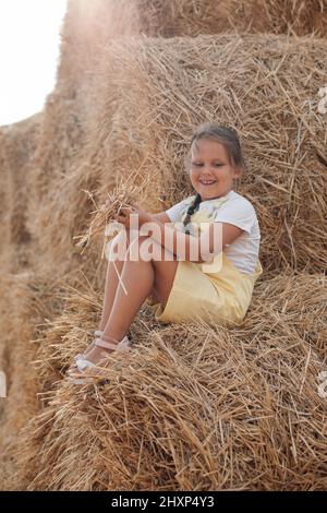 Shining beautiful female child on hayrick smiling looking down and having some hay in hands wearing sundress. Having fun away from city on field full Stock Photo