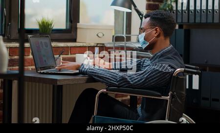 Worker in wheelchair wearing face mask at business job, working on laptop to create financial statistics in disability friendly office. Accountant planning report during covid 19 pandemic. Stock Photo