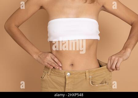 Woman in white bra and large cream colored jeans showing thin waist on flesh background. Healthy eating and body building. Loosing weight by keeping Stock Photo
